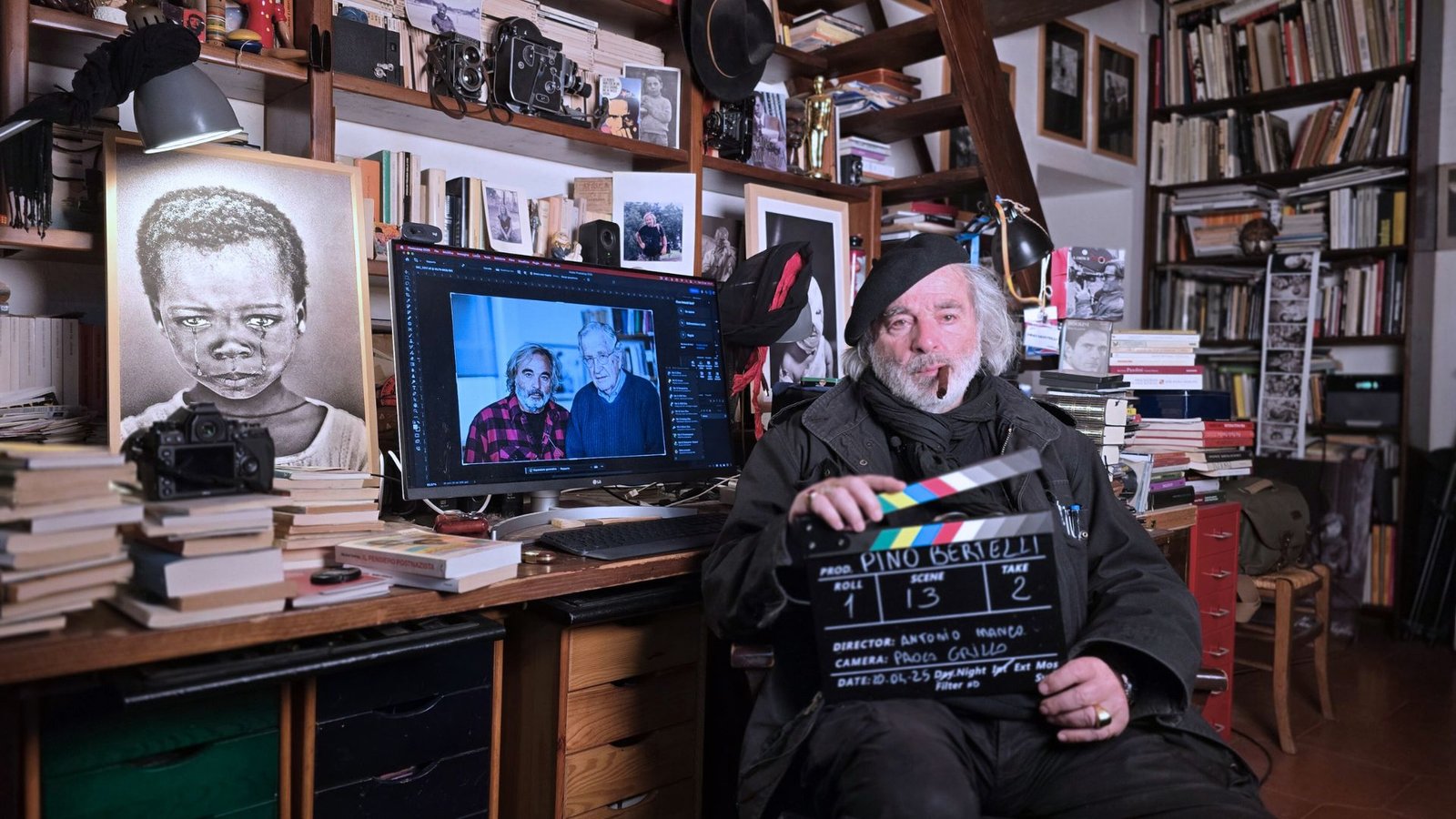 Pino Bertelli seduto con un ciak in mano nel suo studio a Piombino, circondato da libri e fotografie, tra cui una con Noam Chomsky. Backstage del documentario diretto da Antonio Manco.